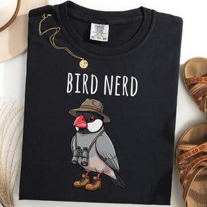Op de afbeelding: Zwart T-shirt met de tekst "BIRD NERD" boven een cartoonvogel met een hoed, verrekijker en laarzen. Een gouden ketting en bruine sandalen zijn ook zichtbaar.