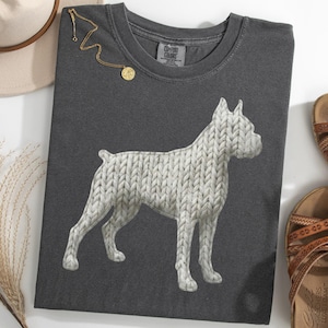 May include: Dark gray t-shirt featuring a white Boxer dog design with a knitted texture. The shirt is surrounded by a beige hat, gold necklace, and brown sandals. A sprig of dried grass is also present.