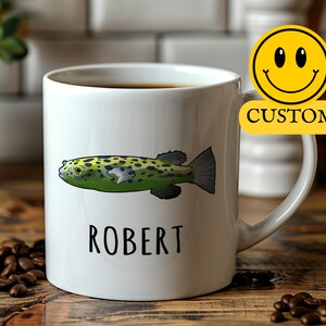 May include: White ceramic coffee mug with a green and black pufferfish illustration and the name "ROBERT" printed in black. The mug is on a wooden surface with coffee beans scattered around it.