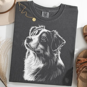 May include: Charcoal grey t-shirt with a detailed white illustration of a dog's face. A gold necklace with a pendant is on the shirt. Brown sandals and a beige hat are also in the image.