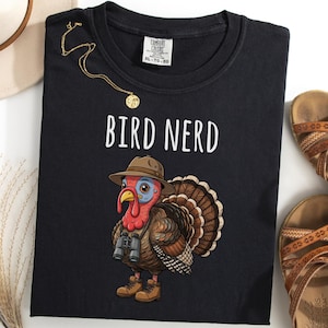 May include: Black t-shirt with the text "BIRD NERD" and a cartoon turkey wearing a hat and binoculars. The shirt is paired with brown sandals and a gold necklace. A beige hat is visible in the upper left corner.