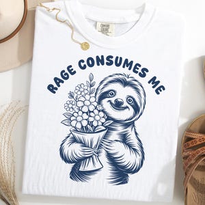 May include: White t-shirt with a navy blue illustration of a smiling sloth holding flowers, with the text "RAGE CONSUMES ME" above the sloth. The shirt is a Comfort Colors brand.