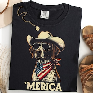 May include: A black t-shirt featuring a graphic of a spotted dog wearing a cowboy hat, sunglasses, and an American flag bandana. Below the dog, the text reads 'MERICA in large, light-colored capital letters. The shirt has a Comfort Colors label.