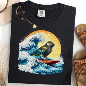 May include: Black t-shirt featuring a parrot surfing a wave with a yellow sun in the background. The shirt has a Comfort Colors tag. A gold necklace, a hat, and sandals are also in the image.