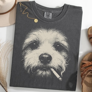 May include: Dark gray t-shirt featuring a detailed illustration of a dog with a cigarette in its mouth. The shirt is accompanied by brown sandals, a gold necklace, and a beige hat.