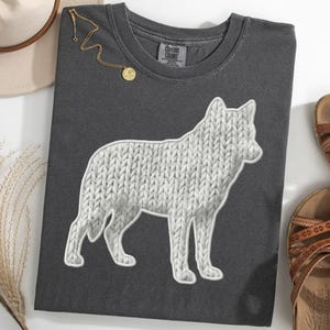 May include: Dark gray t-shirt featuring a white dog silhouette with a knitted texture design. The shirt is accompanied by a gold necklace, a beige hat, and brown sandals. The design is a unique and stylish representation of a dog.