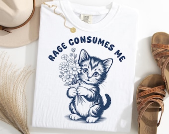 Op de afbeelding: Wit T-shirt met een marineblauwe afbeelding van een katje met bloemen, met de tekst "RAGE CONSUMES ME" erboven. Het shirt is van het merk Comfort Colors.