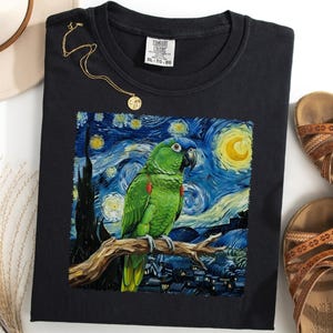 Puede incluir: Camiseta negra con un diseño de loro verde al estilo de 'La noche estrellada' de Van Gogh. También se ven un collar dorado y sandalias marrones. La camiseta es talla XL-TG.