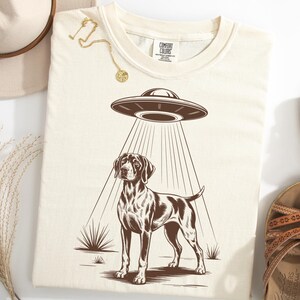 Puede incluir: Camiseta color crema con un gráfico marrón de un perro Pointer siendo abducido por un OVNI. La camiseta está rodeada por un sombrero, sandalias y joyas.