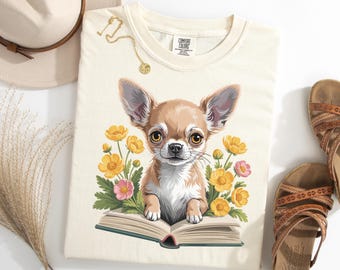 May include: Cream-colored t-shirt featuring a charming chihuahua illustration. The dog is depicted with large ears, brown fur, and expressive eyes, surrounded by yellow and pink flowers, and resting on an open book. A gold necklace and sandals are also visible.