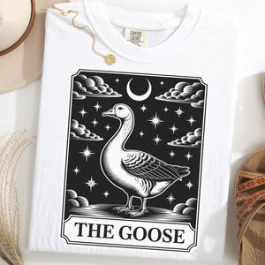 Pode incluir: Camiseta branca com uma ilustração em preto e branco de um ganso, nuvens, estrelas e uma lua crescente. O texto "THE GOOSE" está na parte inferior. A camiseta está dobrada e mostrada com sandálias e um chapéu.