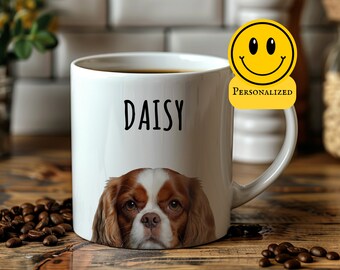 May include: White ceramic coffee mug with a brown and white Cavalier King Charles Spaniel dog peeking out from the bottom. The mug has the name "Daisy" written in black on the front. The mug is sitting on a wooden surface with coffee beans scattered around it.