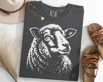 Puede incluir: Camiseta gris oscuro con una ilustración blanca de la cabeza de una oveja. La camiseta está doblada, con un collar dorado y sandalias marrones. Un sombrero beige y hierba seca también están en la imagen.