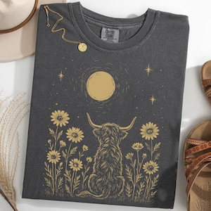 May include: Dark gray t-shirt featuring a golden illustration of a Highland cow, daisies, a full moon, and stars. A gold necklace and brown sandals are also visible. The shirt is folded and ready to wear.