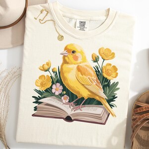 May include: Cream-colored t-shirt featuring a yellow canary perched on an open book, surrounded by yellow and pink flowers. A gold necklace and a beige hat are placed near the shirt, along with brown sandals.