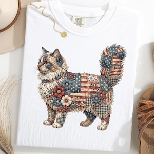 May include: White t-shirt featuring a detailed illustration of a cat with a patchwork design incorporating the American flag, flowers, and patriotic patterns. The cat's fur is a mix of red, white, and blue. The shirt has a Comfort Colors tag.