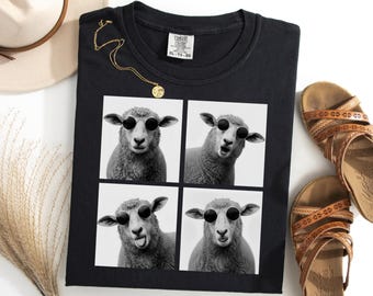Puede incluir: Camiseta negra con una imagen de cuatro paneles de ovejas con gafas de sol. También se ven un collar dorado y sandalias marrones. La camiseta está etiquetada como "Comfort Colors" y es talla XL-TG.