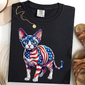 May include: A black t-shirt featuring a graphic of a hairless cat with a red, white, and blue American flag pattern. The shirt has a Comfort Colors label in size XL. The design includes a cat with large ears and eyes, standing in profile.
