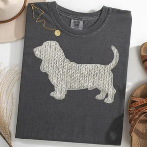 Könnte beinhalten: Dunkelgraues T-Shirt mit einem weißen Basset Hound-Design mit Strickmuster. Eine goldene Halskette mit Anhänger und braune Sandalen sind ebenfalls zu sehen. Das T-Shirt liegt gefaltet auf einer weißen Oberfläche.