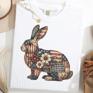 Pode incluir: T-shirt branco com um design de coelho em patchwork. O coelho apresenta estrelas, listras e motivos florais patrióticos em vermelho, branco e azul. A camisa é estilizada com um chapéu, sandálias e um colar.
