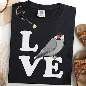 Pode incluir: Camiseta preta com a palavra "LOVE" em branco e uma ilustração de um pássaro cinza. Um colar dourado e sandálias marrons também são visíveis. A camisa tem uma etiqueta Comfort Colors.