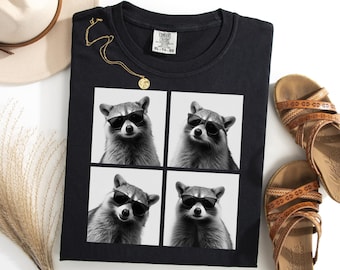 May include: Black t-shirt featuring a four-panel image of a raccoon wearing sunglasses. The shirt is accompanied by a gold necklace, a straw hat, and brown sandals. The shirt has a Comfort Colors tag.