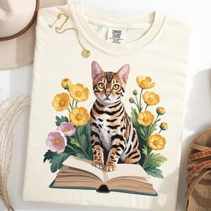 May include: Cream-colored t-shirt featuring a Bengal cat illustration sitting on an open book, surrounded by yellow and pink flowers. The shirt has a Comfort Colors tag. A gold necklace and a hat are visible.