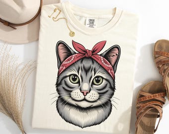 May include: Cream-colored t-shirt featuring a detailed illustration of a cat's face. The cat has gray and white fur, green eyes, and a red bandana tied around its head. The shirt is part of the Comfort Colors collection.