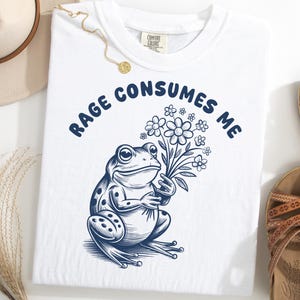 Puede incluir: Camiseta blanca con un gráfico azul marino de una rana sosteniendo flores y el texto "RAGE CONSUMES ME" en arco. La camiseta es de la marca Comfort Colors. La imagen también muestra un sombrero, un collar y sandalias.