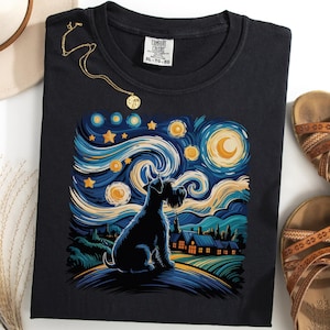 Könnte beinhalten: Schwarzes T-Shirt mit einer stilisierten Illustration eines Hundes vor einem Sternenhimmel, der an den Stil von Van Gogh erinnert. Das Design umfasst einen Mond, Sterne und eine ländliche Landschaft. Eine goldene Halskette und braune Sandalen sind ebenfalls sichtbar.