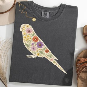 Puede incluir: Camiseta gris oscuro con un diseño floral de pájaro en crema, amarillo, rosa, naranja y morado. La camiseta se combina con un collar dorado, un sombrero beige y sandalias marrones.