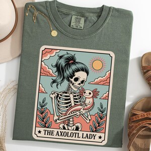 May include: Sage green Comfort Colors t-shirt with a graphic of a skeleton woman holding an axolotl. The graphic is framed with a light blue sky, pink clouds, and a yellow sun. The text "THE AXOLOTL LADY" is at the bottom.