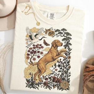 Op de afbeelding: Een crèmekleurig T-shirt met korte mouwen en een grafische print van een bruine hond die op zijn achterpoten staat, een vliegende kraanvogel, een vlinder en diverse bloem- en boomelementen in bruine, zwarte en gouden tinten.