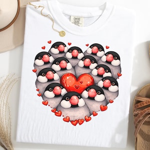 Pode incluir: Camiseta branca com um desenho em forma de coração composto por pássaros de desenho animado com cabeças pretas, corpos cinzentos e bicos rosa. Um grande coração vermelho está no centro, rodeado por corações vermelhos menores. A camiseta está em uma superfície branca.