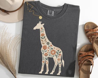 May include: Dark gray t-shirt featuring a floral giraffe design in cream, orange, and green. The shirt is folded, with a gold necklace, a beige hat, and brown sandals visible. A sprig of dried grass is also present.