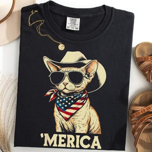 Peut inclure: Un t-shirt noir avec une illustration de chat portant un chapeau de cowboy, des lunettes de soleil noires et un bandana aux couleurs du drapeau américain. Le texte 'MERICA est imprimé en grandes lettres majuscules claires sous le chat.