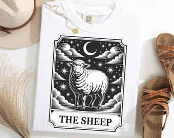 Puede incluir: Camiseta blanca con un gráfico en blanco y negro de una oveja, una luna creciente, estrellas y nubes. El texto "THE SHEEP" se muestra en la parte inferior del gráfico. La camiseta es parte de una colección de ropa.