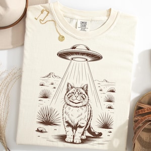 Puede incluir: Camiseta color crema con un gráfico marrón de un gato siendo abducido por un OVNI en un paisaje desértico. La camiseta está rodeada por un sombrero, sandalias y un collar.