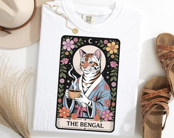 May include: White t-shirt featuring a tarot card design with a Bengal cat illustration. The cat is depicted in a blue kimono holding a cup of coffee, surrounded by flowers. The text "THE BENGAL" is at the bottom.