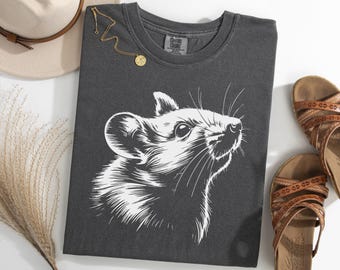 May include: Dark gray t-shirt featuring a detailed white illustration of a rat's face. The shirt is styled with a gold necklace, a beige hat, and brown sandals. The design is a graphic print.