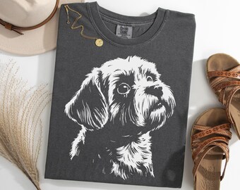 May include: Dark gray t-shirt featuring a white illustration of a dog's face. The shirt is folded and lies on a white surface, next to a hat, sandals, and a gold necklace.