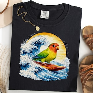 May include: Black t-shirt featuring a colorful parrot surfing a wave with a yellow sun in the background. The shirt is paired with brown sandals, a gold necklace, and a cream-colored hat.