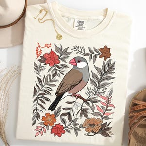 Pode incluir: Camiseta creme com uma ilustração de pássaro. O pássaro é marrom, cinza e rosa, empoleirado em um galho cercado por elementos florais em vermelho, laranja e cinza. Um colar de ouro e sandálias também são visíveis.