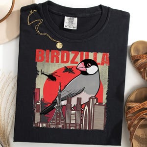 May include: Black t-shirt featuring a graphic of a bird with the word "BIRDZILLA" in red letters. The bird is set against a red sun and city skyline. A gold necklace and brown sandals are also in the image.