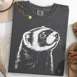 May include: Dark gray t-shirt featuring a white illustration of a ferret's face. The shirt is styled with a gold necklace, a beige hat, and brown sandals. The shirt is made of a soft, comfortable material.