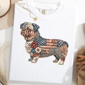 May include: White t-shirt featuring a patriotic pug design. The pug is adorned with a patchwork design incorporating the American flag, floral accents, and a collar. The shirt is styled with a hat, sandals, and a necklace.