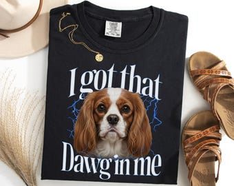 May include: Black t-shirt with the phrase "I got that Dawg in me" and a Cavalier King Charles Spaniel graphic. A gold necklace and brown sandals are also visible.