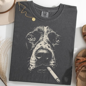 Może przedstawiać: Ciemnoszara koszulka z ilustracją psa trzymającego papierosa. Obraz zawiera brązowe sandały, złoty naszyjnik i beżowy kapelusz. Koszulka jest marki Comfort Colors.