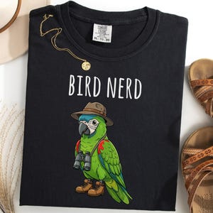 Puede incluir: Camiseta negra con las palabras "BIRD NERD" sobre un loro de dibujos animados con sombrero, prismáticos y botas. También se ven un collar dorado y sandalias marrones.