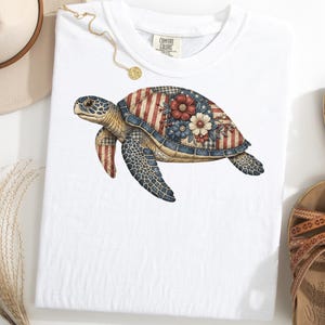 May include: White t-shirt featuring a detailed illustration of a sea turtle. The turtle's shell is decorated with a patriotic design of red stripes, blue floral patterns, and a patchwork effect. The shirt has a Comfort Colors tag.